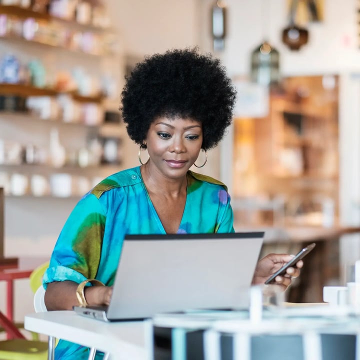 Une femme aux cheveux afro est concentrée sur son ordinateur portable, occupée à travailler ou à étudier dans un environnement lumineux et moderne.