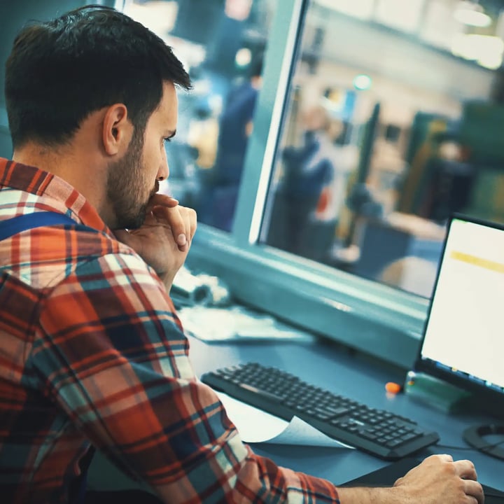 Un homme vêtu d'une chemise à carreaux est assis à un bureau, concentré sur son écran d'ordinateur, entouré d'un espace de travail bien rangé.