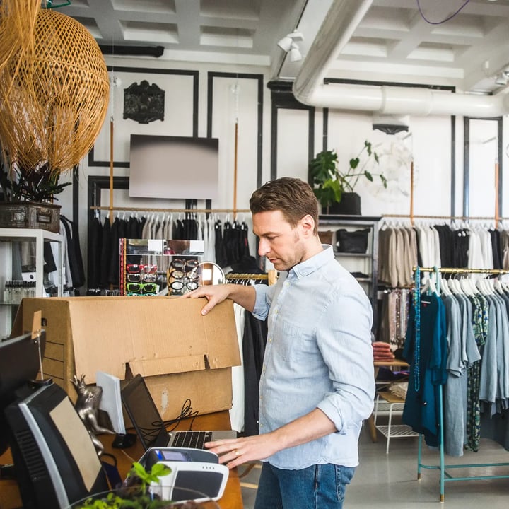 Un homme est concentré sur son ordinateur portable alors qu'il est assis dans un magasin de vêtements, entouré de divers vêtements et présentoirs.