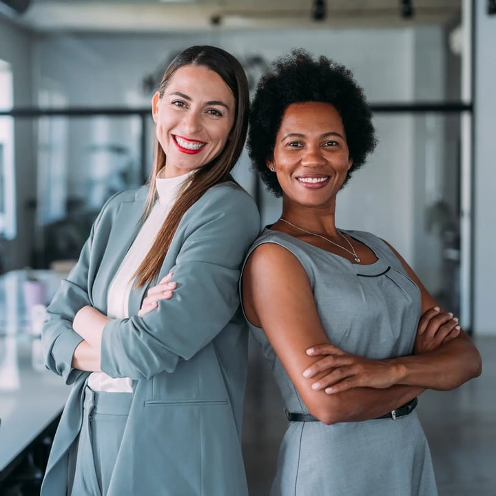 Deux femmes souriantes et posant dos à dos, toutes deux vêtues de tenues grises, dans un bureau moderne.