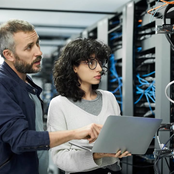 A man and woman working together in a server room, surrounded by servers and technology equipment.