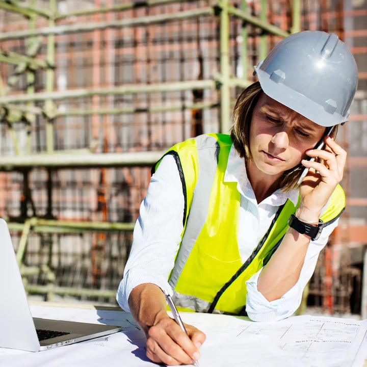 A woman wearing a hard hat and safety vest is engaged in a phone conversation at a construction site.