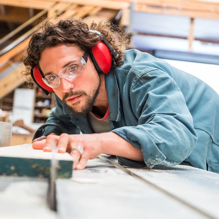 A man wearing ear protection and headphones is focused on his work at a wooden table, ensuring safety and concentration.