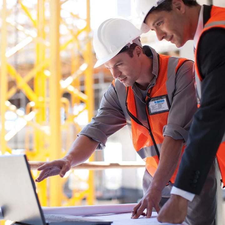 Deux ouvriers de la construction portant des casques de sécurité examinent des plans sur un ordinateur portable sur un chantier de construction.